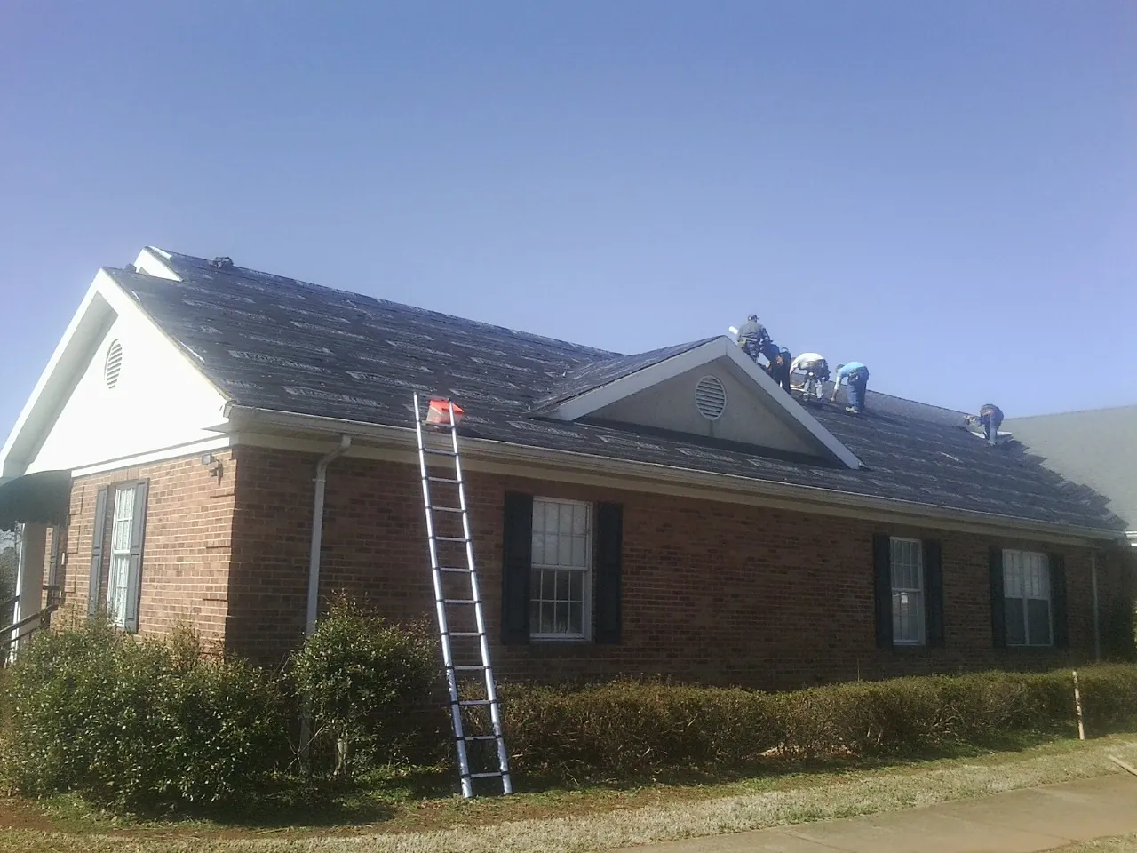 Crew tearing shingles off a brick ranch roof with a ladder set — Emergency Roof Repair work by our crew in Isanti