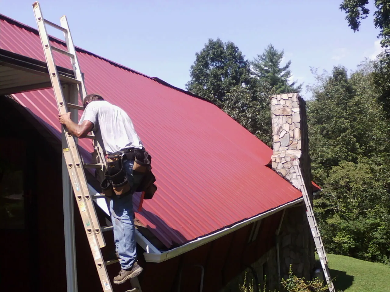 Roofer on a ladder beside a red metal roof and stone chimney — Soffit & Fascia Repair by Roofing Brothers in Isanti